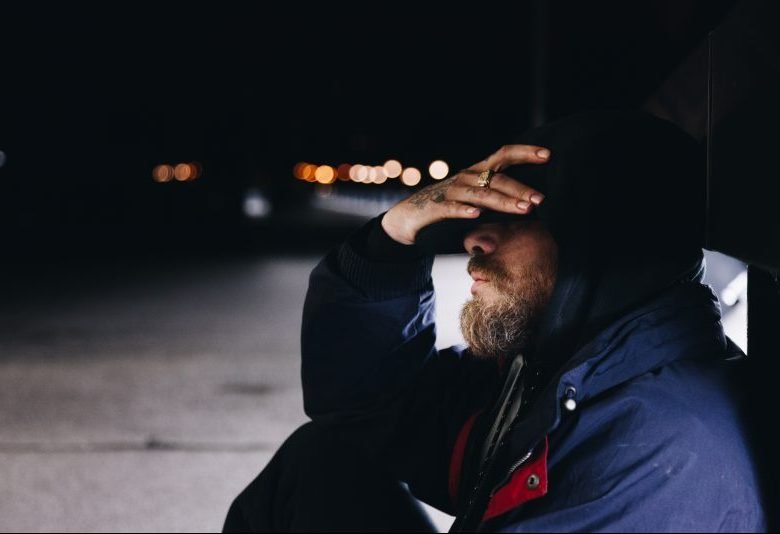 A man sits on a dark sidewalk with his hands over his eyes