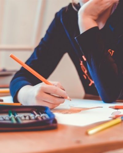 Photo of a student writing with a pencil on a desk scattered with books and classroom materials.