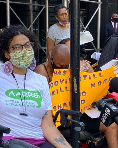 Eman Rimawi, a Black, Native American and Palestinian woman using a wheelchair, wears a tshirt with the Access-a-Ride Reform Group logo and holds up a sign that reads "Don't Kill Us." She is wearing a mask as other masked protestors gather nearby.
