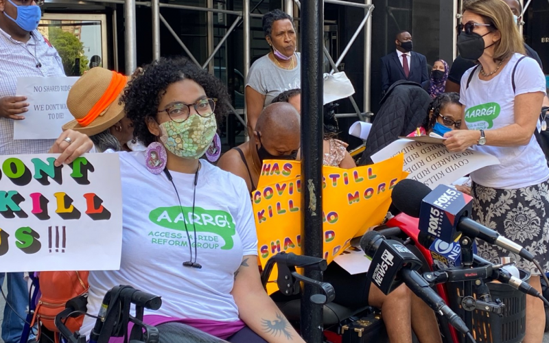 Eman Rimawi, a Black, Native American and Palestinian woman using a wheelchair, wears a tshirt with the Access-a-Ride Reform Group logo and holds up a sign that reads "Don't Kill Us." She is wearing a mask as other masked protestors gather nearby.