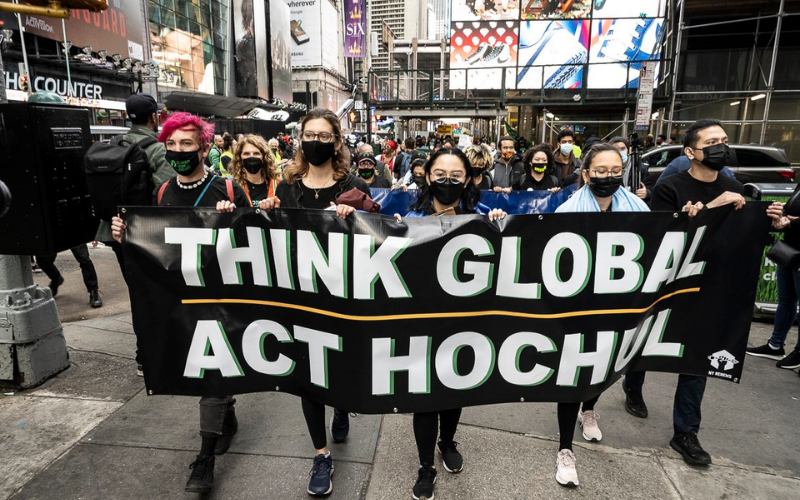 Representatives from NY Renews march through Times Square holding a banner that reads "Think Global, Act Hochul"