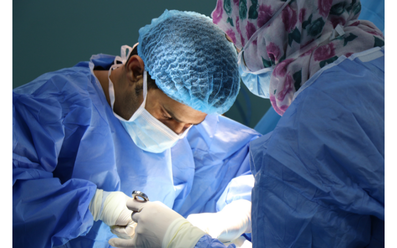 Close-up on a masked surgeon who is operating on a patient that is out of view, with another surgeon's back to us as they look on.