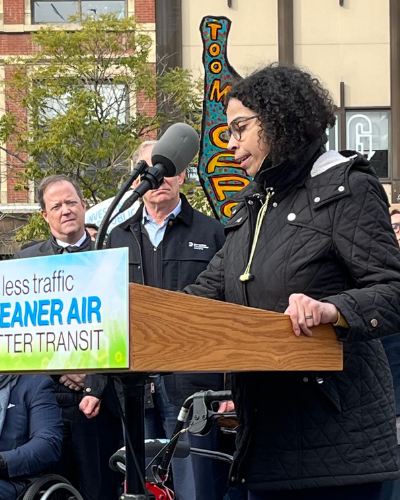 Eman-Rimawi Doster, a woman who is a double amputee with brown hair and wearing a dark coat, stands at the podium and speaks at the Congestion Pricing Rally. Behind her, people hold signs that read "Congestion Pricing Now!"